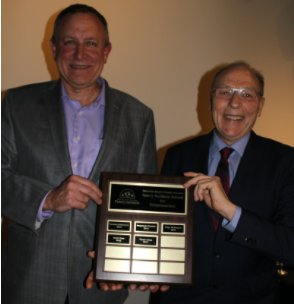 Stuart Cross holding his award up to the camera. He has short, dark hair and wears a grey suit jacket over a blue shirt. He is standing with an older gentleman.